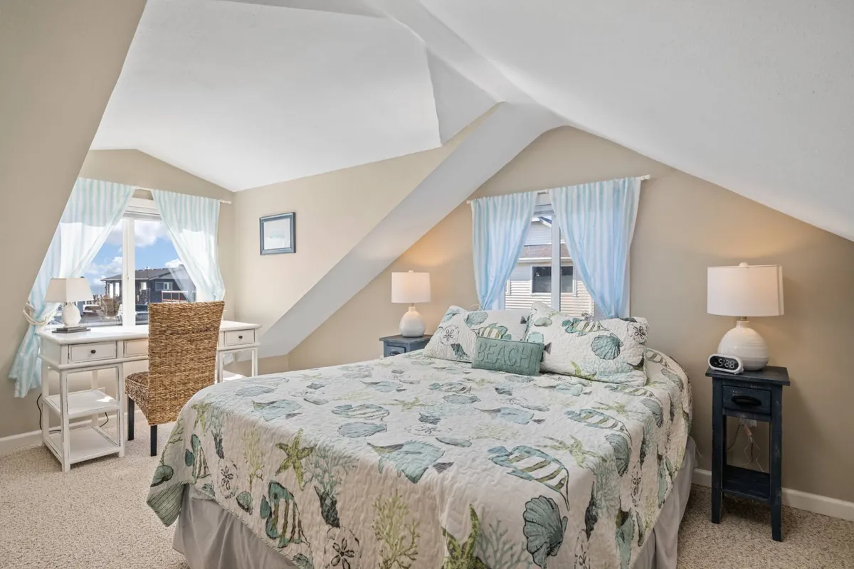 A tranquil upstairs bedroom scene with a bed dressed in beach-themed bedding, a desk and chair positioned by a window with sheer curtains, and calming decor. The workspace offers a glimpse of outdoor light, aligning with the promise of ocean glimpses and a peaceful place to work or relax.