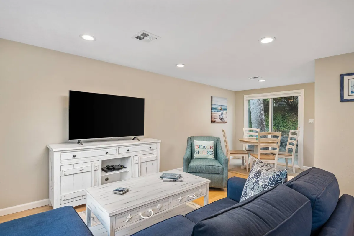 This image shows a bright and inviting living area with a coffee table, television, and beach-themed decor. The space gently flows into the dining area, with large windows that suggest an airy, open atmosphere and subtle glimpses of the outdoors.