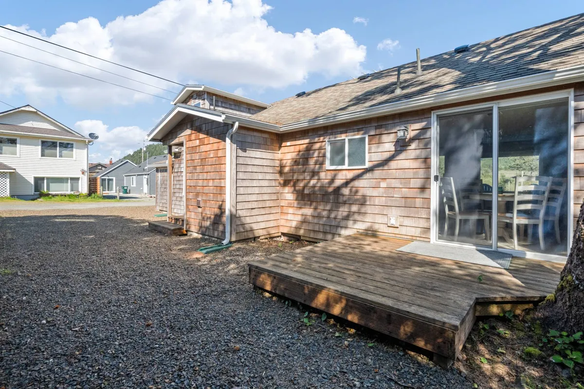A wooden back deck with a sliding glass door, surrounded by neighboring houses and minimal landscaping. The deck offers seating and a relaxed outdoor setting ideal for both people and pets to unwind, dine, or enjoy the fresh air.