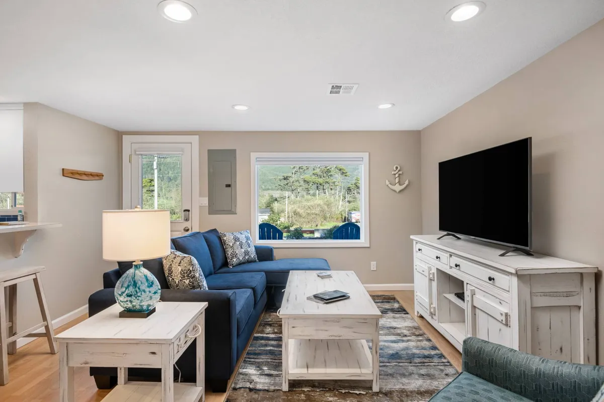 A bright and inviting living room featuring a cozy blue sofa, a crisp white coffee table, a large television, and a generous window that fills the space with natural light and offers glimpses of the scenery just outside. The space feels welcoming, perfect for relaxing with loved ones after a day at the beach.