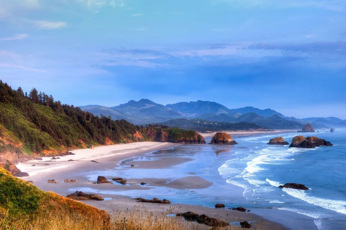 A scenic view of distant mountains, unique rock formations, a sandy beach, and ocean waves. The image captures the beauty and variety of the Oregon coast, suggesting adventure and exploration just beyond the doorstep.