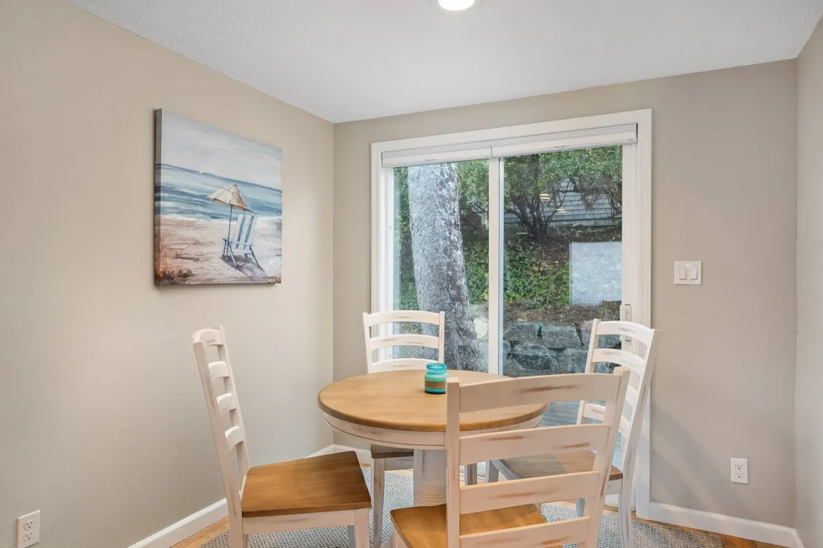 A sunlit dining area featuring a round wooden table with white chairs, beach-themed artwork, and a large window offering a view. The space feels airy and ideal for both casual breakfasts and memorable family dinners.