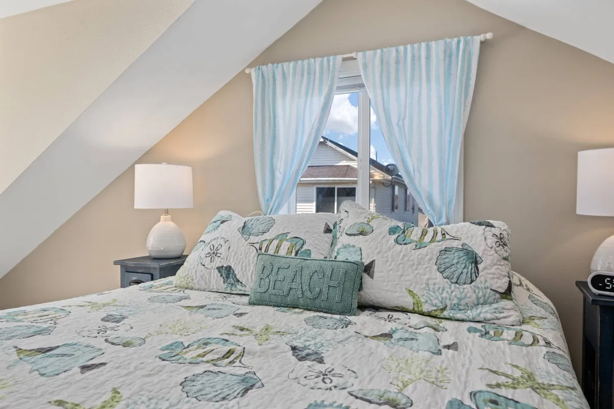A tranquil bedroom featuring a bed with a patterned quilt, a nightstand with a lamp, and a window draped with light curtains. The view from the window hints at the ocean beyond, bathing the room in gentle light and evoking a sense of calm.