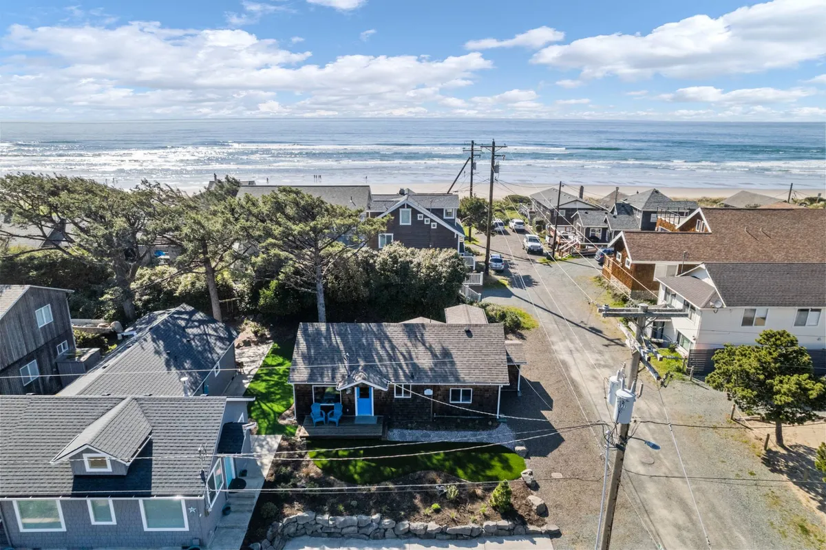 A beachfront view showcasing quaint houses nestled among lush greenery, with the wide sandy shore and gentle ocean waves just steps away. The scene invites thoughts of leisurely beachcombing and relaxing walks along the water's edge, with the promise of beautiful sunsets on the horizon.