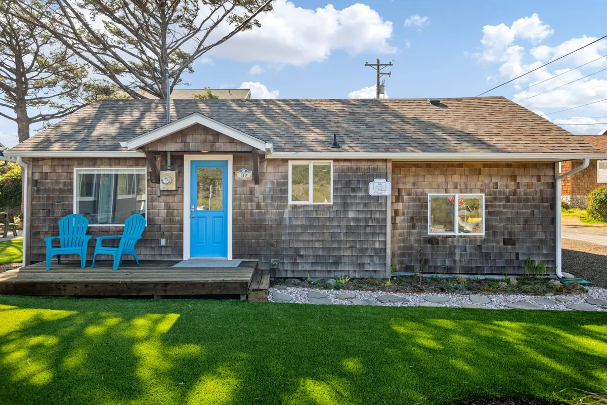 A charming front patio with adirondack chairs and a blue front door, set against wooden siding. The space feels inviting and is ideal for relaxing with a pet after a walk, with easy access to the main entrance and a view of the garden.