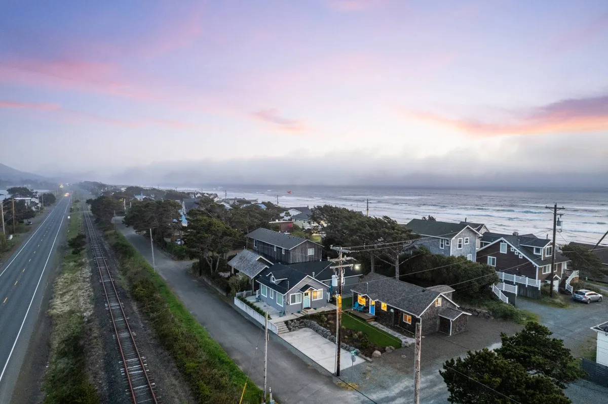A wide coastal view featuring coastal houses, the ocean, a sunset sky, a road, and visible train tracks. The image evokes the unique sense of place, hinting at the seasonal railway experience mentioned in the section, with scenic train tracks running close to the property.