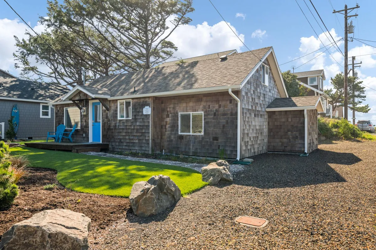 An exterior shot showcasing a blue door, adirondack chairs, landscaped lawn, and rock features. The image captures an inviting and comfortable outdoor gathering space with lush surroundings, suggesting opportunities for alfresco dining, relaxing, or unwinding with a pet.