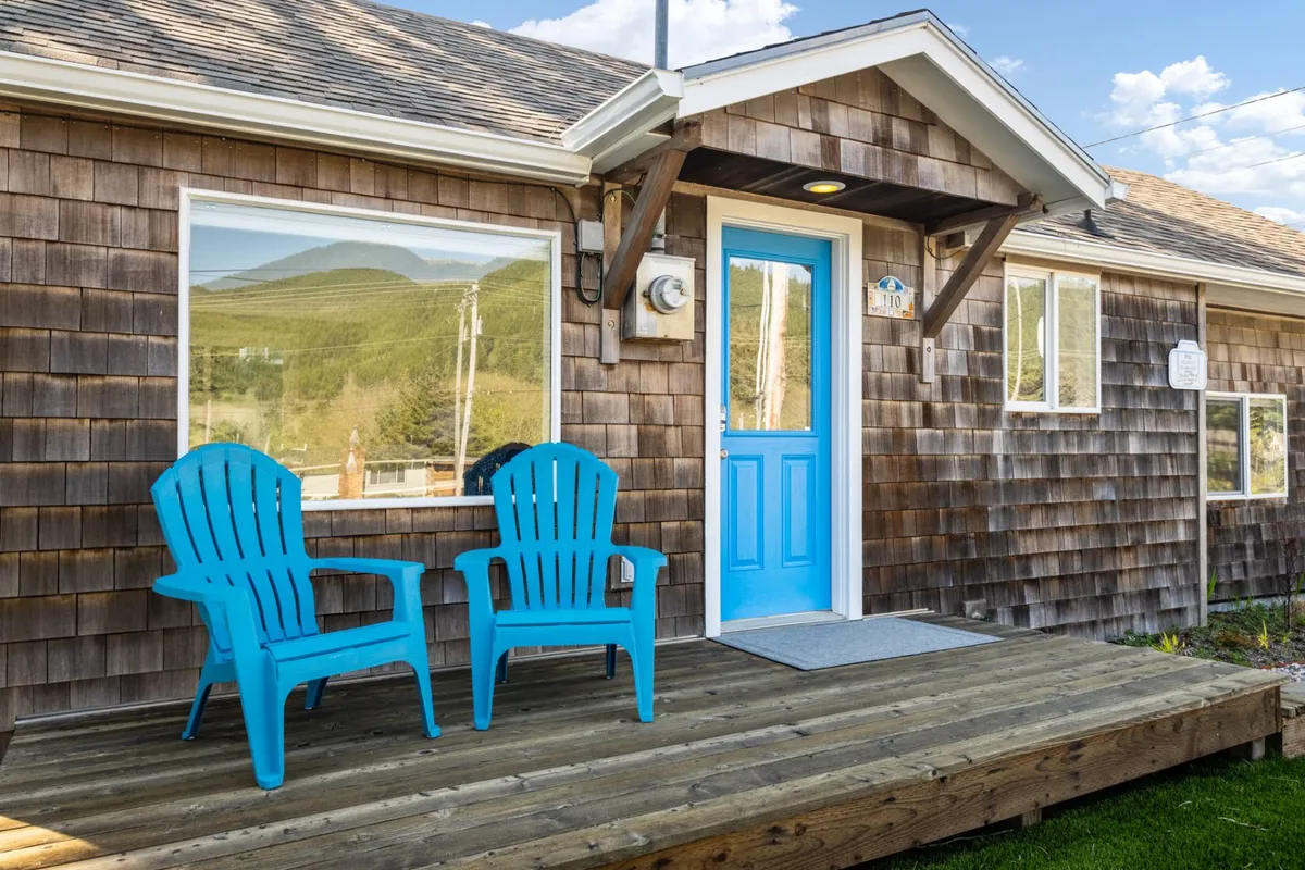 This exterior shot features turquoise chairs arranged invitingly on a wooden deck, with a blue front door that adds a cheerful touch. The scene feels warm and welcoming, suggesting a perfect spot to relax and watch the world—or a vintage train—go by.