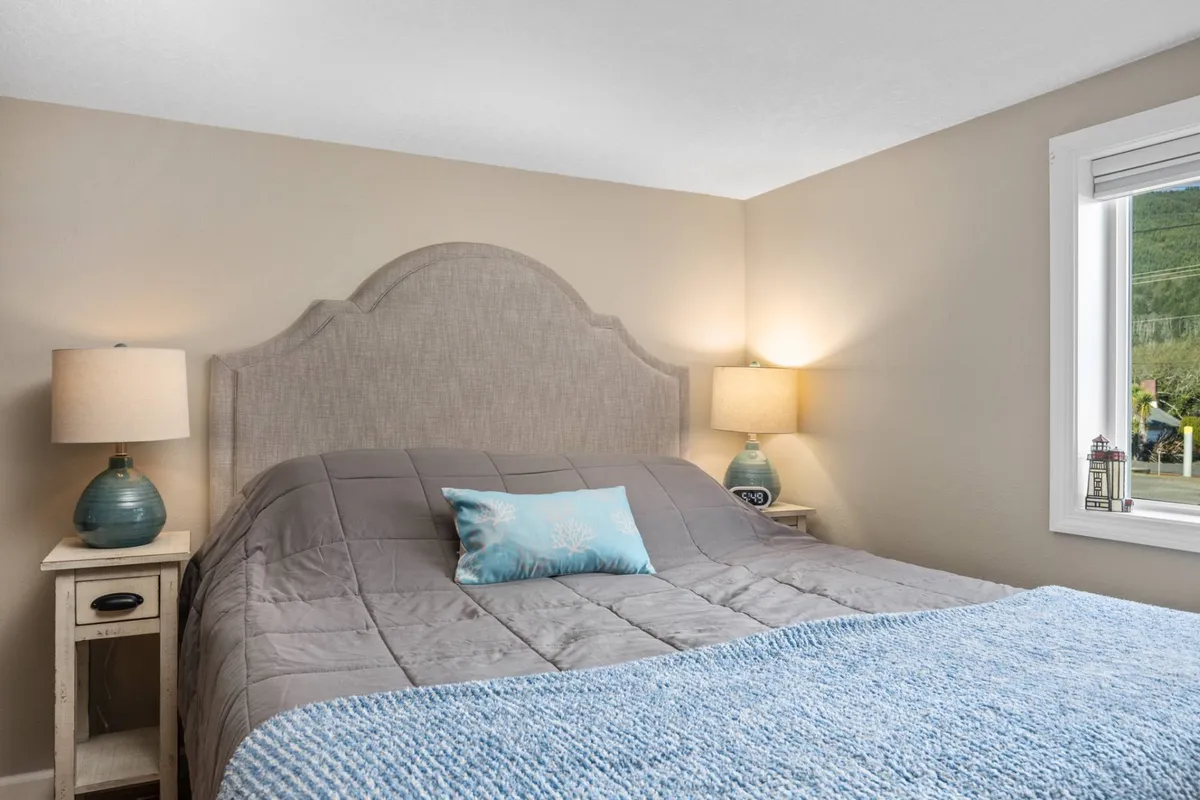 A serene bedroom scene focusing on a comfortable bed with a decorative headboard and accent pillows. Natural light pours in through a nearby window, and a bedside lamp adds to the calming vibe, emphasizing rest and relaxation.
