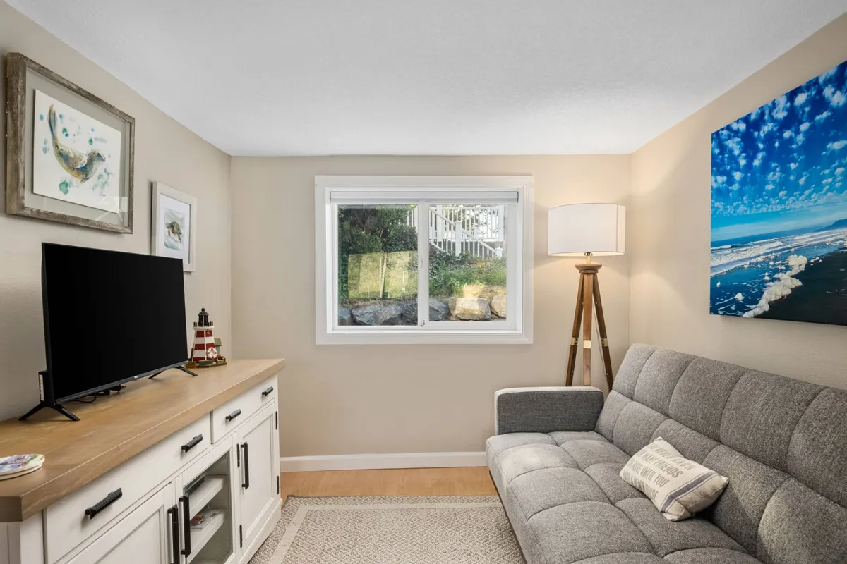 A cozy main-level living area featuring a gray sofa, TV, lamp, and a window with partial ocean artwork. The layout is open and inviting, with carpeted flooring and ample space for lounging. The decor is neutral and calming, making it a comfortable area for both guests and their pets to relax together after a day outdoors.