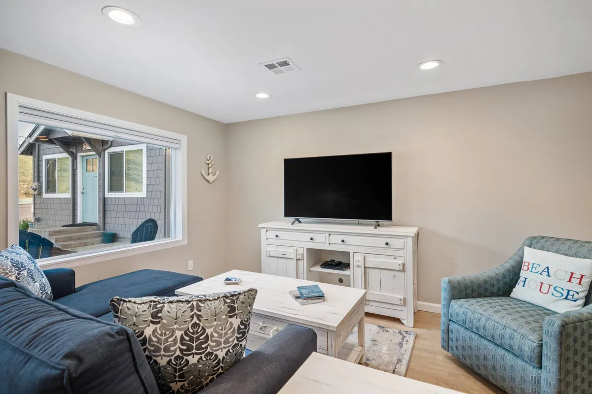 A cozy living area with a blue sofa, coffee table, television, accent chair with a beach-themed cushion, and a large window bringing in sunlight. The space is decorated in a relaxed, coastal style and invites guests to unwind or gather for a movie night.